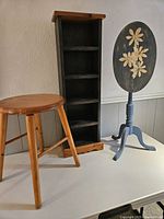 Three wood furniture pieces: spindle stool, tall black shelf, and floral oval drop leaf table