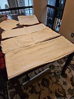 Four large vintage linen tablecloths spread out on a table showing delicate damask and embroidered patterns along with napkins in background.