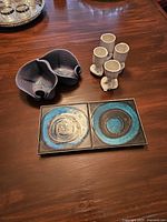 Overview of all items on wooden table: two-piece candy and nut dish, four small boot-shaped liquor vessels, ceramic trivet with blue and brown circles