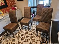 Four rattan wrapped Parsons style chairs arranged on a patterned area rug in a dining room, showing front and side views with natural light from nearby windows.