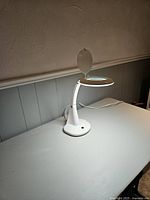 White magnifying lamp on a table, lamp switched off showing the hinged lens cover closed.