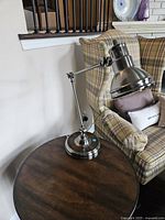Articulating metal table lamp placed on a wooden side table next to a plaid armchair.