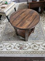 Angle view of full circular end table showing wooden top, lower shelf, and vertical supports, placed on patterned carpet next to upholstered chair.