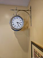 Front view of double-sided wall clock mounted on wall bracket, showing one clock face with Roman numerals and black hands.