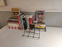 Photo of the full collection of 23 cookbooks arranged next to the black iron book stand on a shelf.