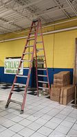 Full view of the 12-foot orange fiberglass ladder standing open in a room with tiled floor and yellow wall, next to some stacked boxes.