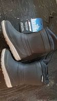 Pair of black Baffin insulated winter boots with original tags attached, laid on a dark surface showing side profile and sole