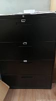Front view of tall black metal filing cabinet with four drawers and minor surface scratches.