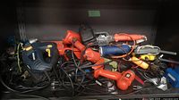 Multiple electric power tools and accessories piled together on a shelf, including Black & Decker drills, a blue saw, and cords.
