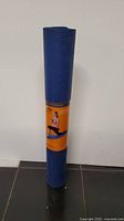 Front angled view of the rolled blue yoga mat with an orange label showing a woman sitting stretching.