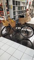 Full view of the black Daymak Paris electric bicycle showing the front wicker basket, the frame, and wheels on a tiled floor indoors with storage boxes in the background.