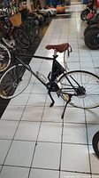 Full side view of black Daymak Vermont 36V electric bicycle with brown synthetic saddle, water bottle holder, showing the overall bike condition and setup in a showroom with other vehicles around.