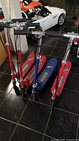 Four children’s electric scooters lined up, three in red and one in blue with visible black handles and chrome stems, showing overall condition and color variations.