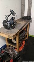 Wooden workbench with mounted metal vise and bench grinder sitting on top. Various tools stored underneath and around workbench.