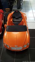 Front view of orange Bentley ride-on toy car showing detailed grille, headlights, and quilted black seat.