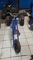 Front view of blue trike showing front wheel, pedal arms, decorative plate and handlebar