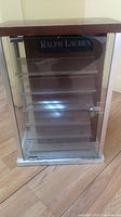 Front view of rectangular display case with brown top, silver base and glass front door with lock showing several horizontal glass shelves inside. Ralph Lauren branding visible on back panel.