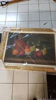 Front view of framed still life painting covered in protective plastic, showing a reddish jug with various fruits on a dark background.