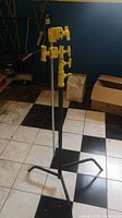 Full view of the yellow metal photography lighting stand showing the adjustable pole and base with three legs on black and white tiled floor.