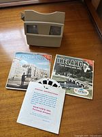 View of the beige View-Master toy viewer sitting on a wooden surface, alongside two slide packets labeled England and Ireland, and the instructional insert visible in the foreground.
