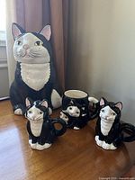 Photo of the full ceramic set showing a large cat cookie jar, two cat salt and pepper shakers, and a cat mug, all black and white with pink details on ears.