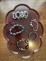 Photo of five assorted bracelets on a wooden clover-shaped table, including various styles and bead types such as black teardrop stones, matte black beads, amber and black beads, and purple bead chain bracelet.