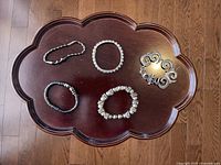 Top-down photo of five silver-colored bracelets on a dark wooden tray, showing varied styles including beads, charms, and scroll cuff.