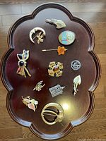 Top-view photo showing 11 assorted vintage brooches on a dark wooden tray arranged in a scattered layout.