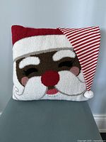 Front view of crochet pillow with a Santa Claus face showing closed eyes, red nose, white beard, and red hat with white trim.