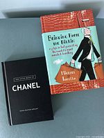 Pair of hardcover books on a gray surface: one book with a black cover titled 'The Little Book of Chanel', the other with an orange textured cover and illustrated blue jacket titled 'Bringing Home the Birkin'.