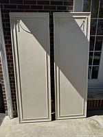 Two tall French Provincial style wooden doors leaning against a brick wall next to a white framed glass door, showing carved garlands and panel details.