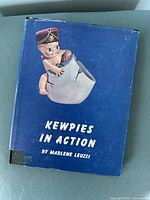 Front cover of 'Kewpies in Action' book showing blue cover with Kewpie doll hatching from an egg and text title and author
