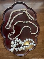 Three necklaces displayed on a wooden flower-shaped table including rhinestone, pastel braided silver chain, and pearl bead cluster necklaces.