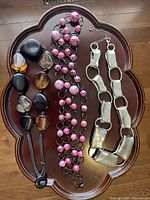 Three necklaces displayed on brown scallop-edged tray: large bead leather necklace, pink bead chain necklace, and silver-tone link chain necklace.