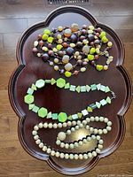 Photo showing three necklaces arranged on a dark wooden decorative tray: multi-colored beaded necklace with green, brown, cream beads; green geometric bead necklace; cream round bead necklace.