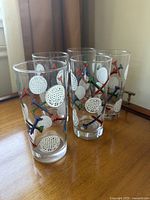 Set of five clear drinking glasses with golf ball and tee designs arranged on a wooden surface near a window.