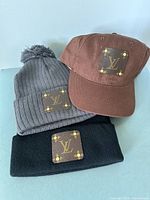 Image showing three headwear items: brown cap, grey toque with pom-pom, and black toque, all with brown Louis Vuitton style patches