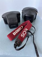 Two vintage camera cases and one red Canon EOS strap displayed on a flat surface.