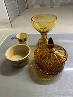 Two amber glass pieces: a covered dish with finial and a goblet or compote dish; alongside two basket-style pottery pieces: a small pitcher and a round bowl with floral motifs.