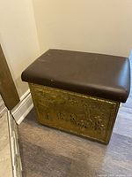 View of brass kindling box with padded seat lid from one side showing embossed scenic design