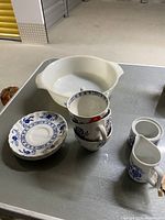 Photo showing the Fire King white casserole dish with blue floral decoration, blue and white teacup stacked with 50% off sticker, matching saucers, milk jug and sugar pot missing lid.
