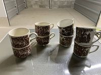 Two sets of brown ironstone coffee mugs, each set stacked in pairs, showing pastoral and village scenes in detailed brown transferware style.
