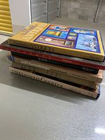 Stack of six coffee table books placed in a storage or garage area, showing visible titles and different thicknesses of books.