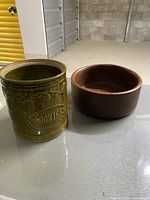 Photo showing green ceramic cookie jar with embossed 'COOKIES' text and round brown wooden bowl side by side on a concrete floor.