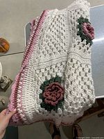 Folded white crochet blanket with pink flowers and green leaves detail, showing open stitch pattern and finished pink border.