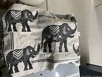 Photo of a rectangular fabric rug with black elephant designs on a cream background, folded and placed on a grey surface.