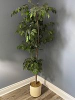 Photo of tall faux tree with green leaves in a white pot positioned in a corner against a grey wall and wooden floor