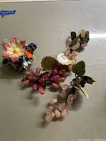 Top view of three glass grape clusters in pink tones with bronze metal leaves alongside a colorful ceramic flower frog with painted flowers.