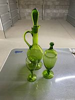 Green glass decanter with handle and stopper, green glass candy dish with lid, and stemmed cup on table viewed from front