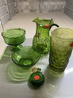 Green glassware lot displayed on metallic table including two candy dishes, pitcher, decorative balloon, and vase.